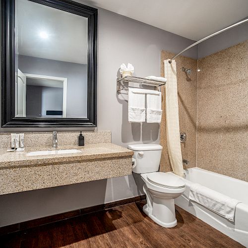 A well-lit bathroom with a granite countertop, large mirror, toilet, towels, and a bathtub with a shower curtain on a dark wooden floor.