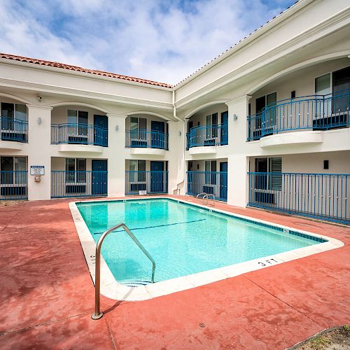 This image shows a small, clean swimming pool surrounded by a red pool deck, situated in the courtyard of a two-story white building with blue railings.