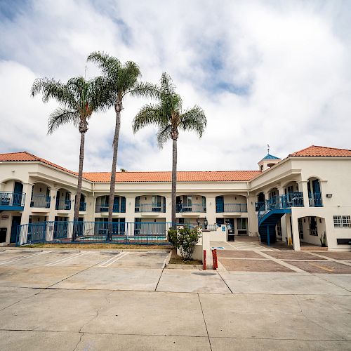 An outdoor view of a two-story building with palm trees in front, blue railings and staircases, and a spacious concrete area.