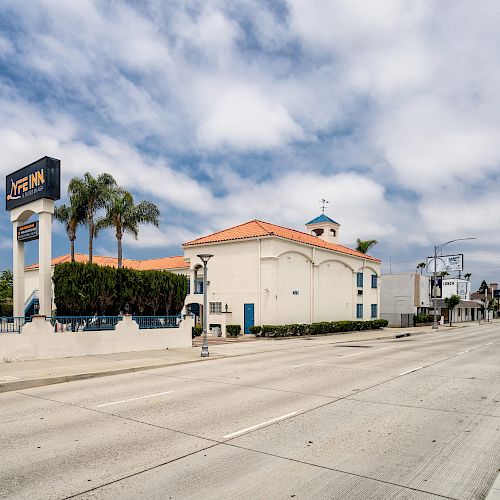 A quiet street with a hotel and a church, featuring palm trees and a blue sky with clouds, is depicted in this image.