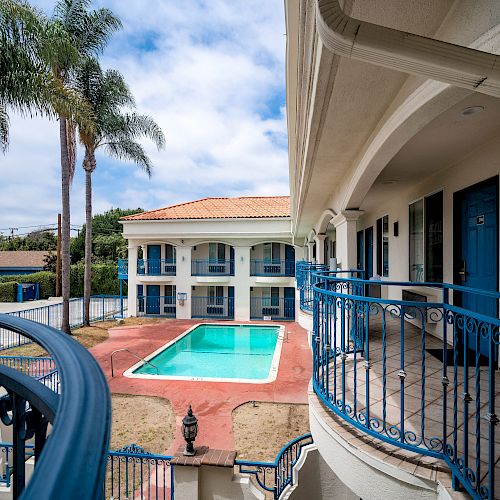 The image depicts an outdoor area of a hotel or motel with a swimming pool surrounded by balconies, featuring palm trees and blue skies in the background.