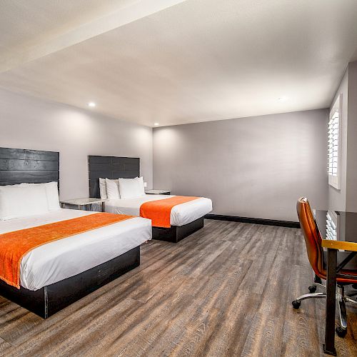 A modern hotel room with two double beds, orange throws, a wall-mounted TV, a wooden desk, a chair, and gray walls.