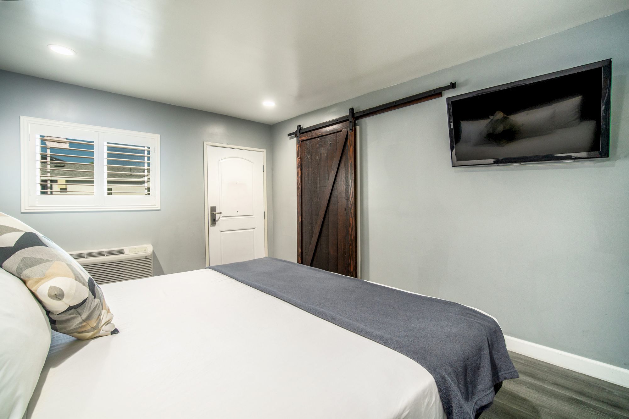 A modern bedroom with a made bed, grey walls, a wall-mounted TV, a wooden barn door, white door and window with shutters, and an air conditioning unit.