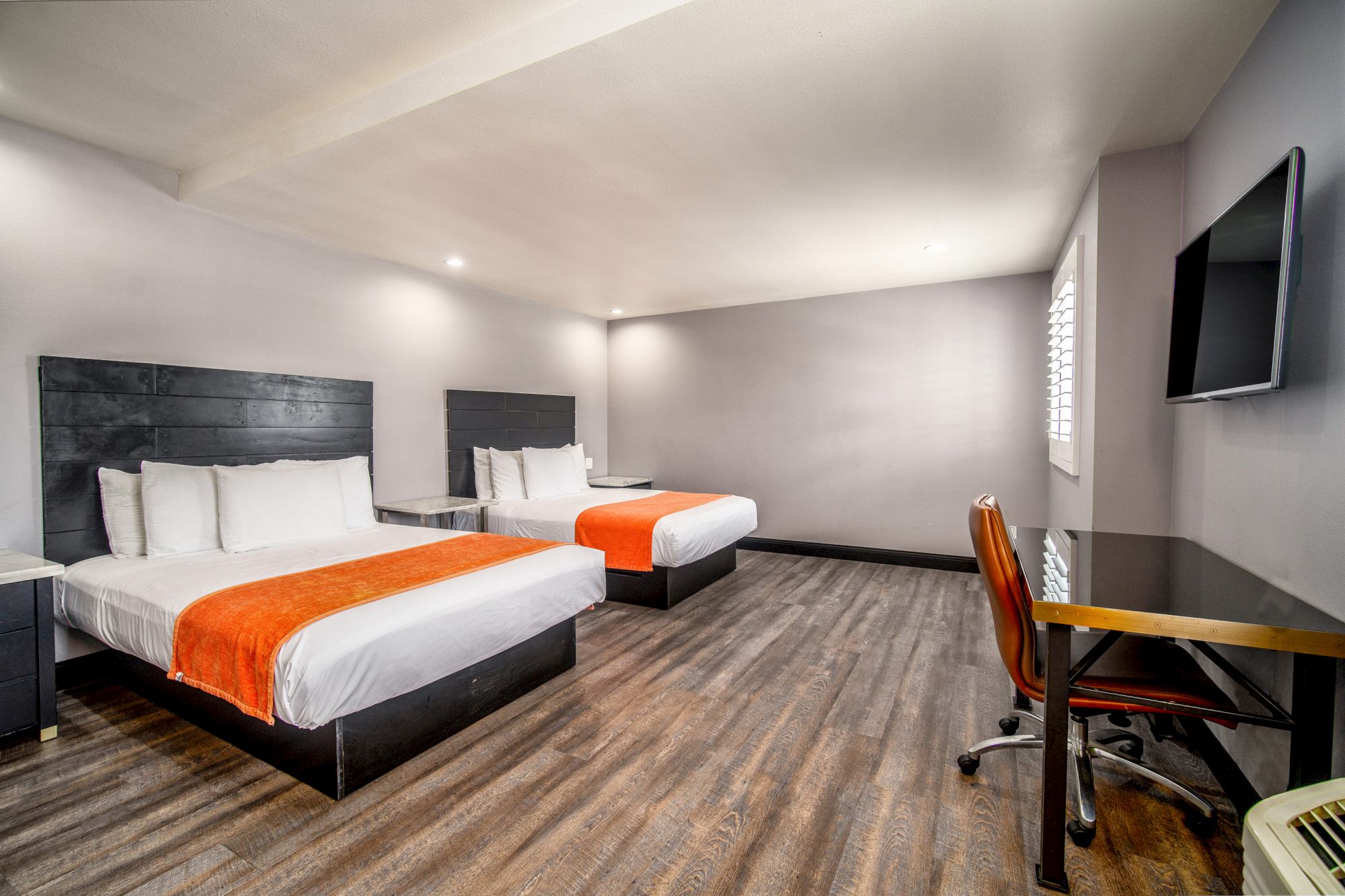 A modern hotel room with two double beds, orange throws, a desk, chair, large TV, wood flooring, and minimalist decor, brightly lit.