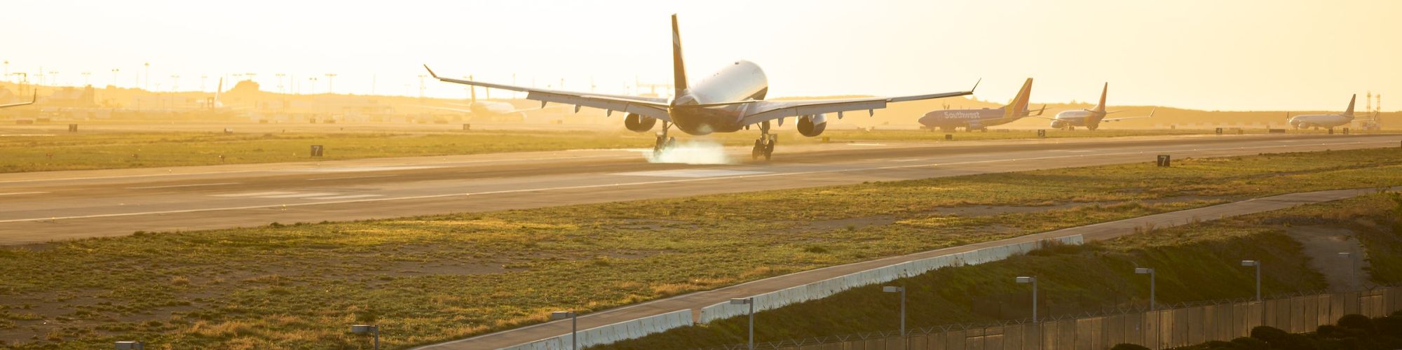 An airplane is landing on a runway during sunset, with other planes in the background and a fence in the foreground.