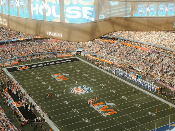 A large football stadium filled with spectators, featuring a game in progress on a marked field, with "RAMS HOUSE" displayed on a prominent screen.
