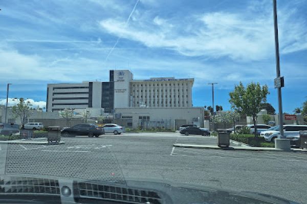 This image shows a large hospital building with a large parking lot and several cars parked in the foreground under a clear, blue sky.