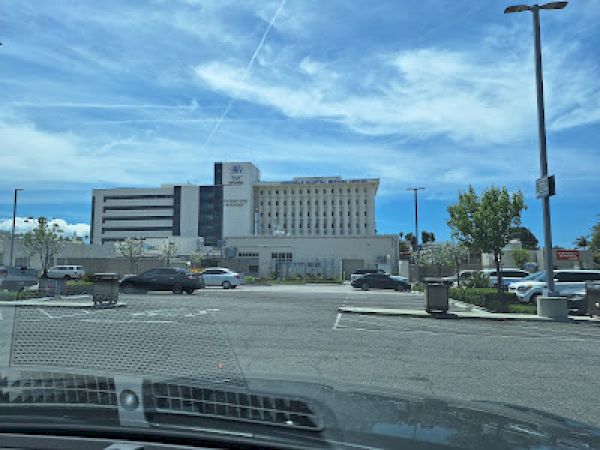 This image shows a large hospital building with a large parking lot and several cars parked in the foreground under a clear, blue sky.