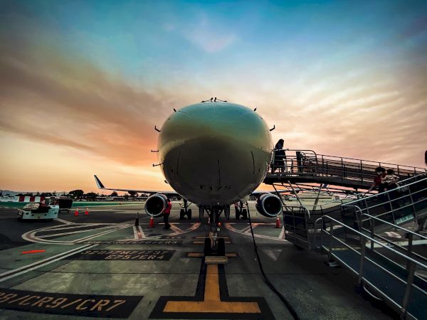 An airplane is parked on the tarmac at an airport during sunset, with a jet bridge connected to it and a clear sky in the background.