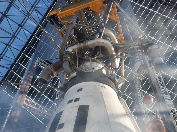 The image shows a view of a rocket engine or spacecraft component inside a large metal framework, with visible pipes and structural elements.