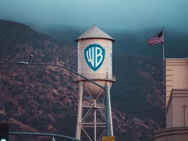 The image shows a Warner Bros. water tower with a large "WB" logo, an American flag, mountains in the background, and a street sign reading "Olive Ave."