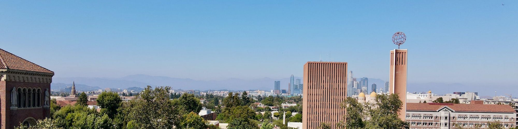 This image shows a green, tree-lined campus with tall buildings, likely part of a university, with a city skyline visible in the distant background.