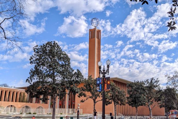 The image shows a brick building with a tall bell tower, trees, a grassy area, and a street with bike lanes. The sky is partly cloudy.