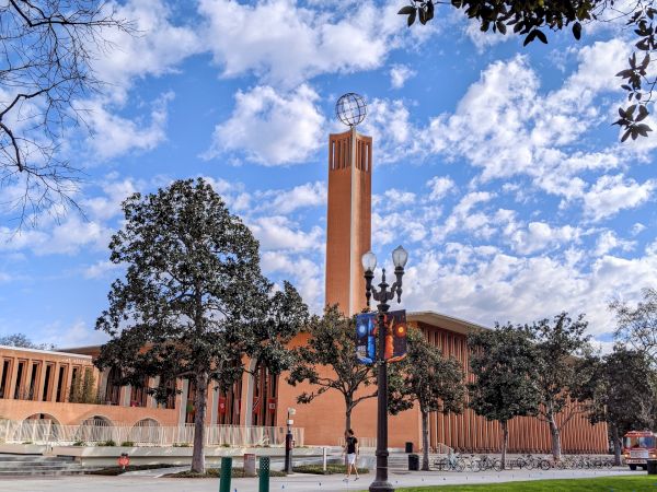 The image shows a brick building with a tall bell tower, trees, a grassy area, and a street with bike lanes. The sky is partly cloudy.