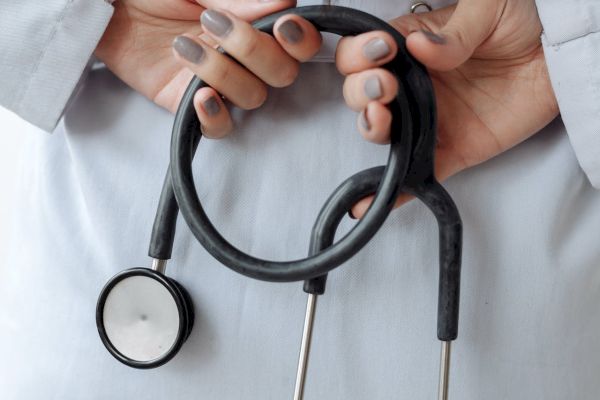 A person in a white coat holds a stethoscope with both hands behind their back. The focus is on the hands and the stethoscope.