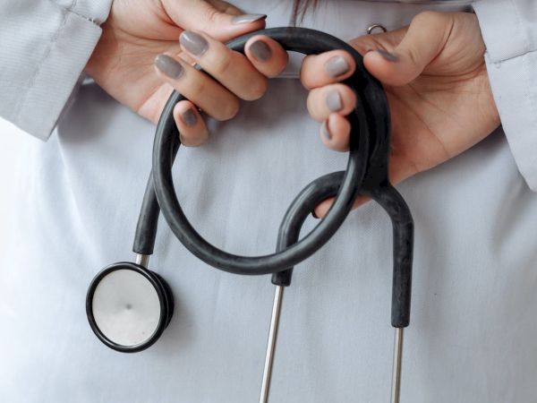 A person in a white coat holds a stethoscope with both hands behind their back. The focus is on the hands and the stethoscope.