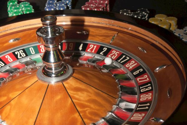 A roulette wheel is in focus with people placing bets using various colored chips on a casino table in the background.