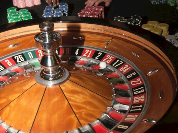 A roulette wheel is in focus with people placing bets using various colored chips on a casino table in the background.