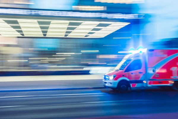 The image shows a red ambulance with flashing blue lights speeding through an urban area at night, with motion blur capturing its swift movement.