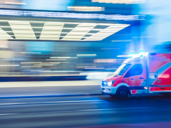 The image shows a red ambulance with flashing blue lights speeding through an urban area at night, with motion blur capturing its swift movement.