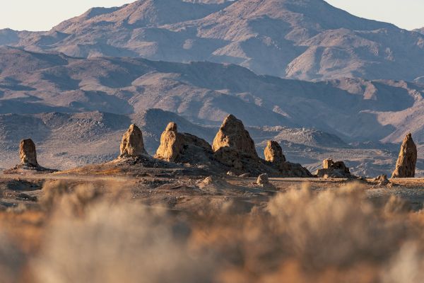 The image shows a dry, rocky landscape with tall rock formations in the foreground and a range of mountains in the background under a clear sky.