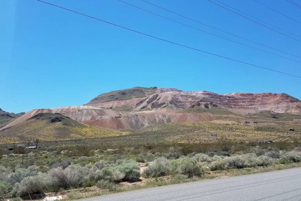 The image shows a paved road leading towards a hillside with various shades of brown and green, possibly indicating a mining site. Power lines run above.