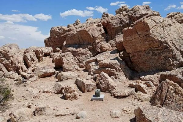 The image shows a rocky desert landscape with large boulders under a clear blue sky. A small monument or marker is present near the center.