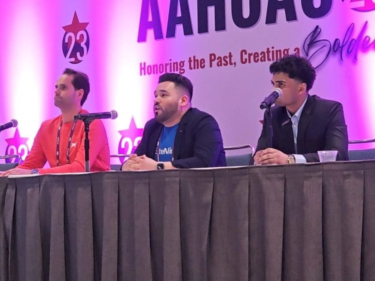 Three people are sitting at a table with microphones, participating in a panel discussion at an AAHOA event.