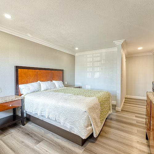 A modern bedroom with a large bed, bedside table, wall-mounted TV, and microwave on a counter. Neutral colors and wood flooring are featured.