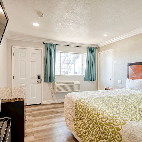 The image shows a neat hotel room with a bed, a TV on the wall, a microwave, and an air conditioner under a window with green curtains.