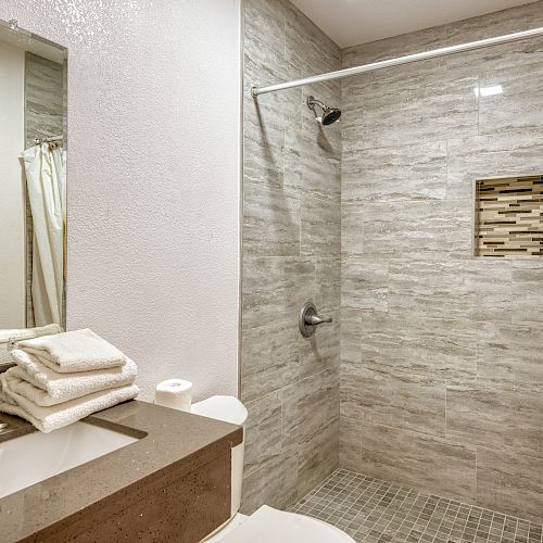 The image shows a modern bathroom with a walk-in shower, a large mirror, a sink with a faucet, and neatly folded towels on the countertop.