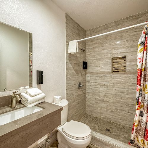 The image shows a modern bathroom with a sink, mirror, toilet, and walk-in shower featuring a colorful curtain and neatly folded towels.