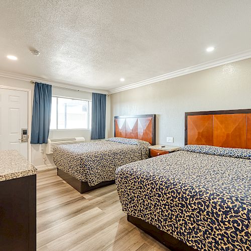 A hotel room with two beds, a TV, a desk, an air conditioner under the window, and blue curtains. The room has wood flooring and beige walls.