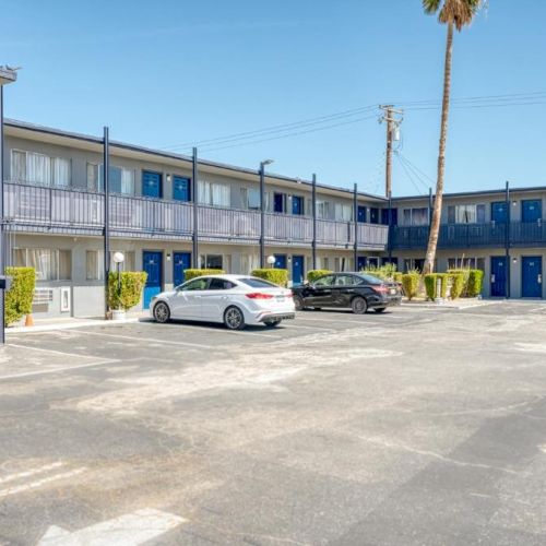 The image shows the parking area of a two-story motel with blue doors, white walls, and several parked cars with a clear blue sky.