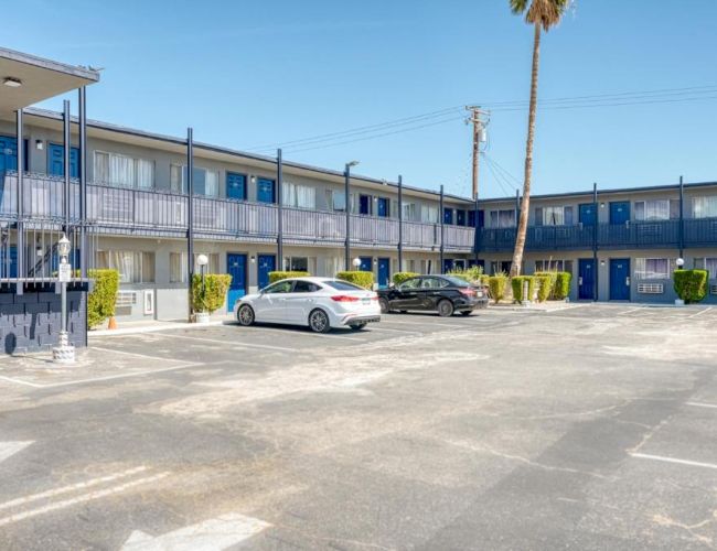 The image shows the parking area of a two-story motel with blue doors, white walls, and several parked cars with a clear blue sky.