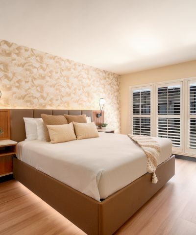 The image shows a modern bedroom with a large bed, decorative wallpaper, bedside lamps, potted plants, hardwood floor, and shuttered windows.