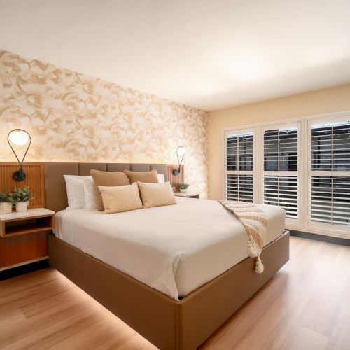 The image shows a modern bedroom with a large bed, decorative wallpaper, bedside lamps, potted plants, hardwood floor, and shuttered windows.