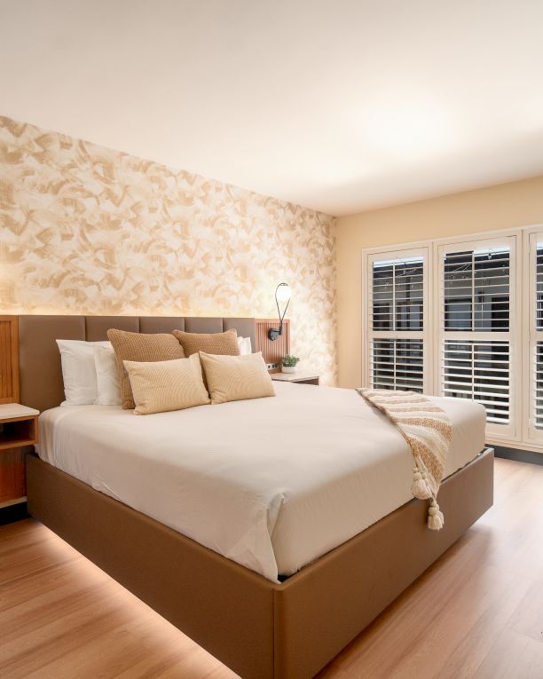 The image shows a modern bedroom with a large bed, decorative wallpaper, bedside lamps, potted plants, hardwood floor, and shuttered windows.