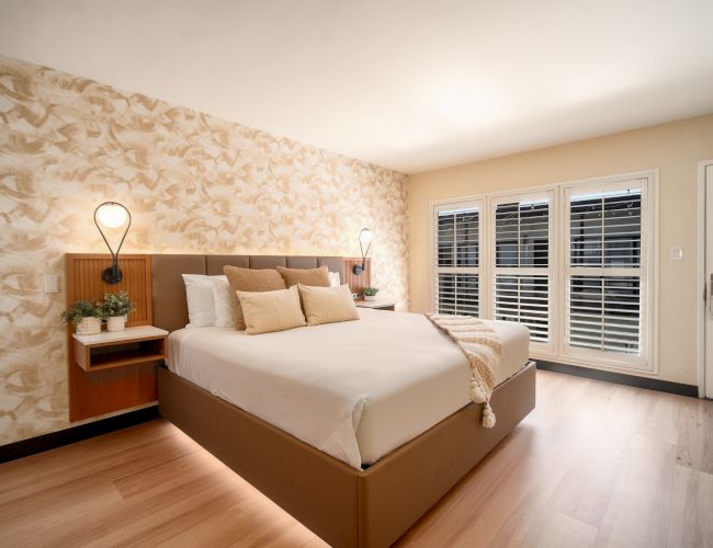 The image shows a modern bedroom with a large bed, decorative wallpaper, bedside lamps, potted plants, hardwood floor, and shuttered windows.