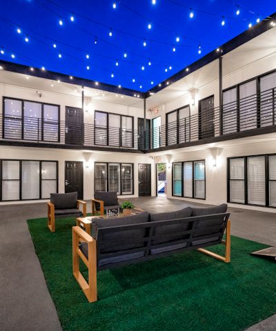 A modern courtyard with seating on artificial grass, surrounded by building units and string lights overhead in the evening.