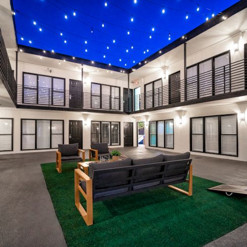 A modern courtyard with seating on artificial grass, surrounded by building units and string lights overhead in the evening.