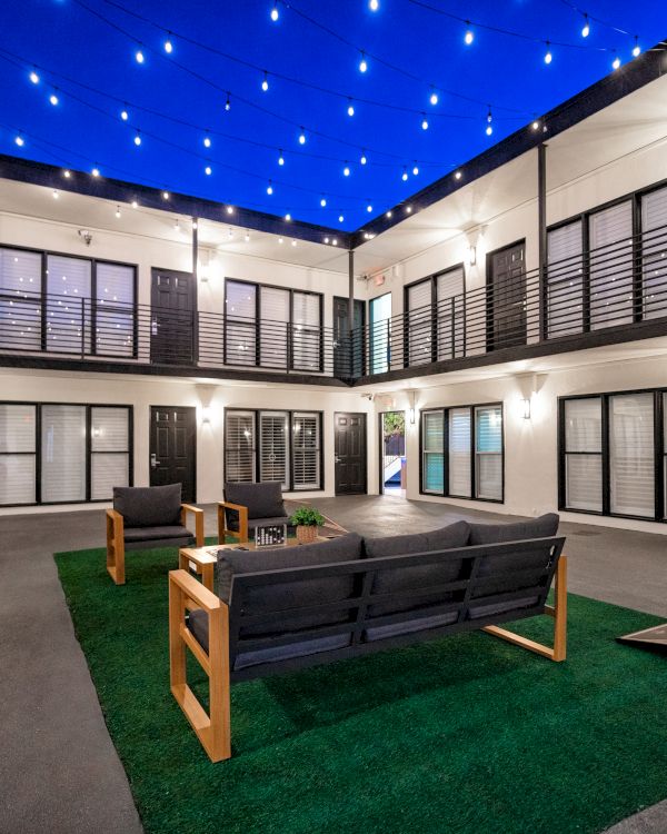 A modern courtyard with seating on artificial grass, surrounded by building units and string lights overhead in the evening.