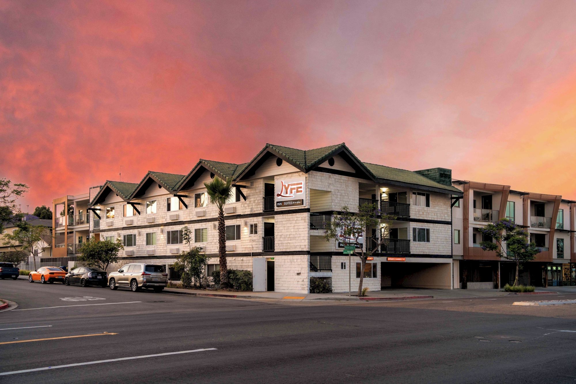 A multi-story apartment building with beige siding sits along a wide street at dusk, the sky glowing orange-pink behind it.
