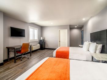 The image shows a modern hotel room with two double beds, a flat-screen TV, a work desk with a chair, and minimalistic decor.