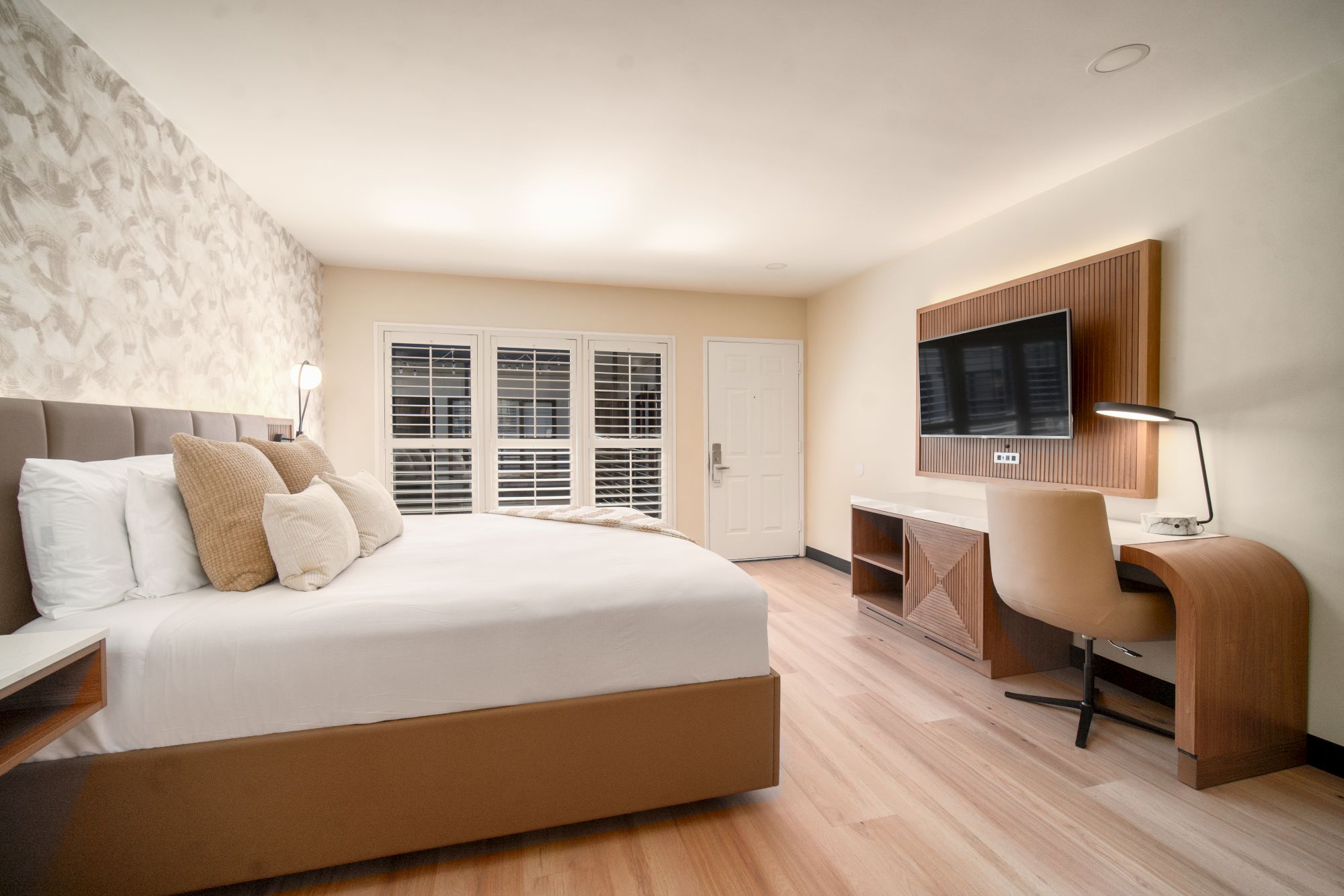 A modern, cozy hotel room features a king-sized bed with multiple pillows, a desk with a chair, a flat-screen TV, and large windows with shutters.