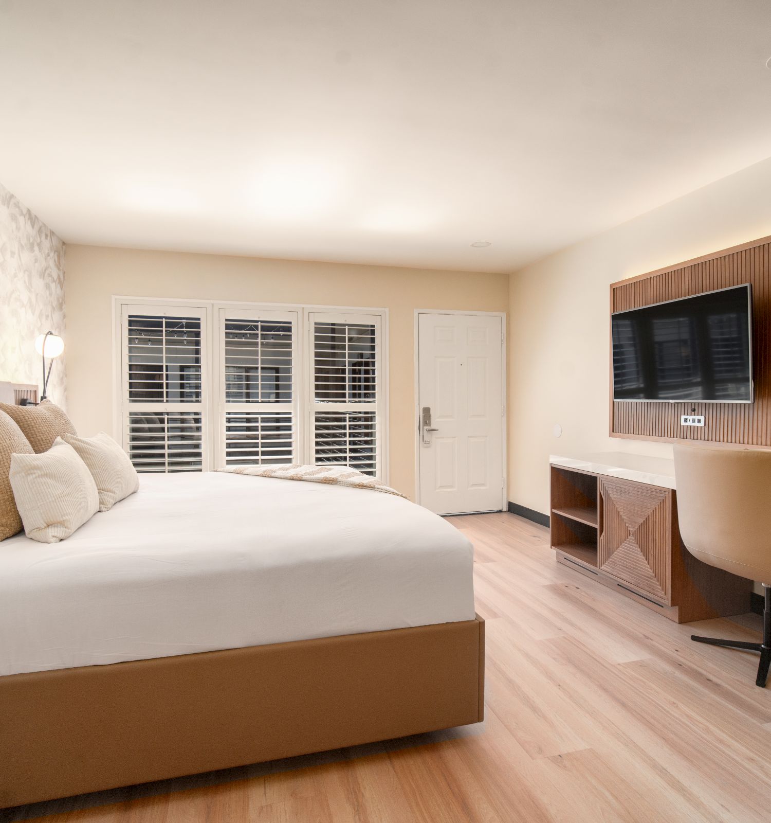 A modern hotel room features a large bed, bedside tables, a flat-screen TV on a wall-mounted desk, a chair, and a window with shutters.