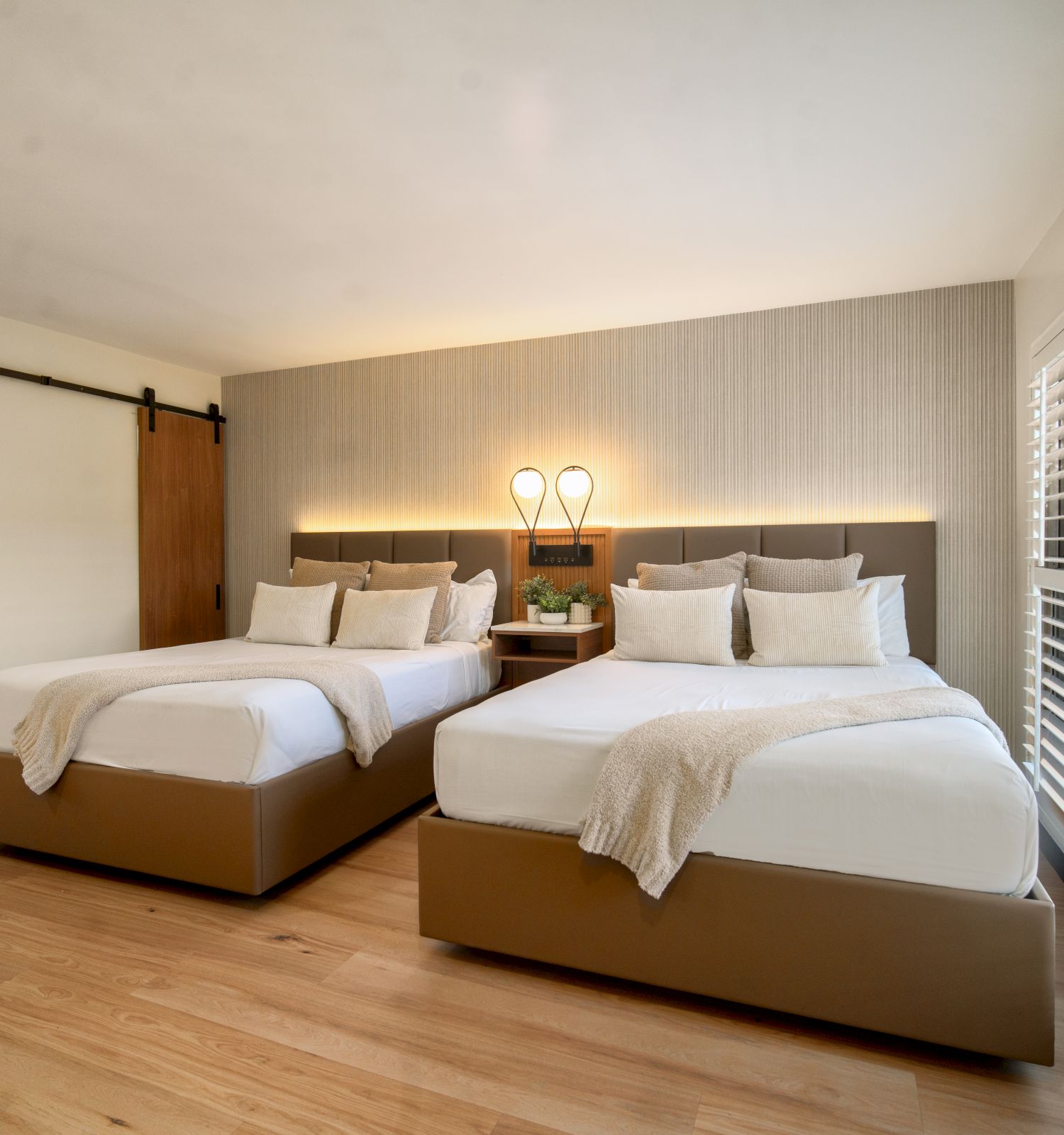 A bedroom with two beds, a chair, wooden flooring, a sliding barn door, and ample natural light from the large window with white shutters.