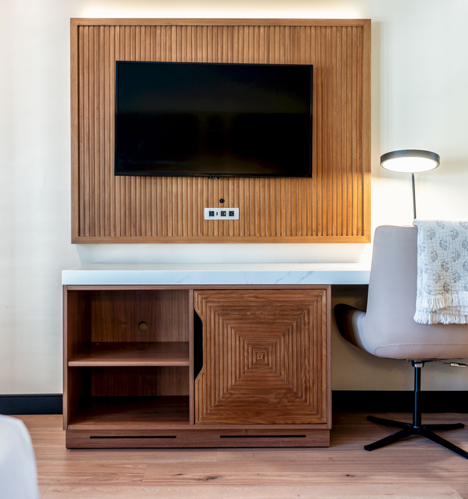 A hotel room features a TV on a wooden panel, a desk with a lamp, a chair, and a blanket draped over the chair.