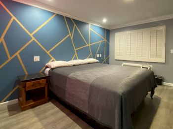 The image shows a modern bedroom with a geometric accent wall, a bed with gray bedding, wooden nightstands, and recessed lighting, always ending the sentence.