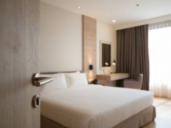 A neatly arranged hotel room with a bed, desk, and chair, viewed through an open door with soft lighting from a window.